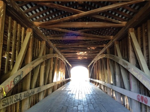 A wooden covered bridge interior with beams and graffiti, leading to a bright light at the end.