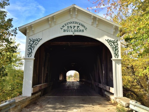 A historic covered bridge with "M. Kennedy & Sons, 1877, Builders" inscribed above the entrance, surrounded by trees.