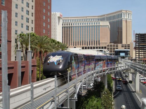 A monorail travels above a busy street, with hotels and palm trees in the background under a clear blue sky.