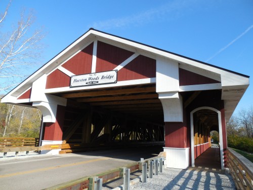 A red and white covered bridge with a sign reading "Hawston Woods Bridge," surrounded by trees and blue sky.