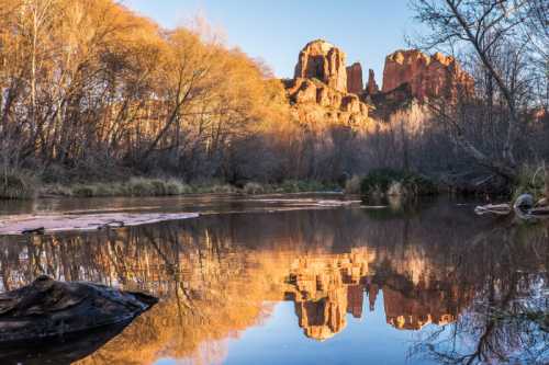 A serene landscape featuring red rock formations reflected in a calm river, surrounded by autumn trees.