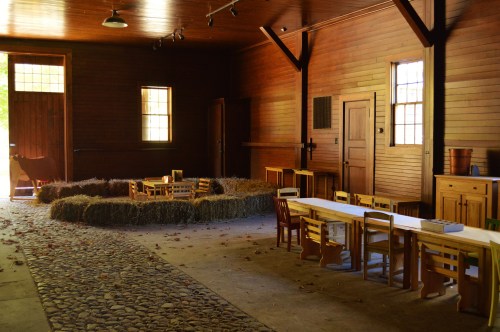A rustic room with wooden walls, a stone floor, and a hay circle, featuring small tables and chairs for children.