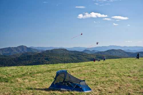 A blue tent on a grassy hill with people flying kites against a backdrop of mountains and a clear blue sky.