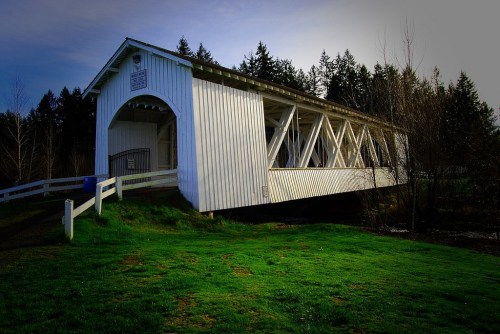 A white covered bridge spans a grassy area, surrounded by trees under a clear sky.