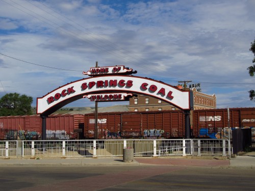 Sign reading "Home of Rock Springs Coal" with freight train cars in the background under a cloudy sky.