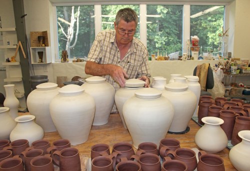 A potter carefully arranges unglazed ceramic pots and jars in a workshop filled with pottery.