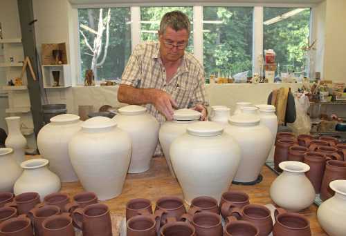 A potter carefully arranges unglazed ceramic pots and jars in a workshop filled with pottery.