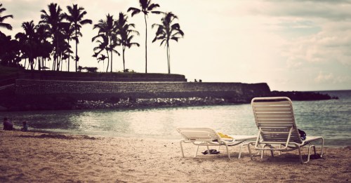 Two empty lounge chairs on a sandy beach, with palm trees and a calm ocean in the background.