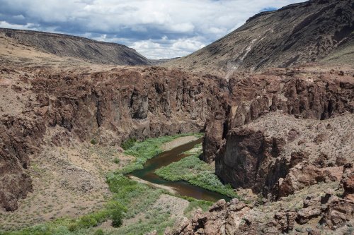 A winding river flows through a rugged canyon surrounded by rocky cliffs and sparse vegetation under a cloudy sky.