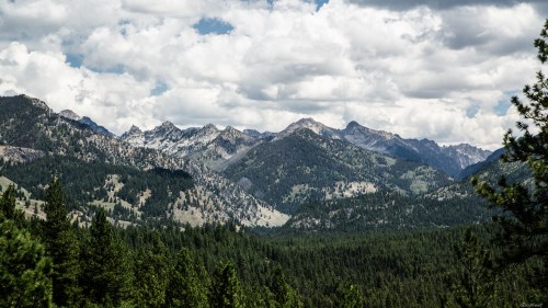 A panoramic view of rugged mountains under a cloudy sky, surrounded by lush green forests.