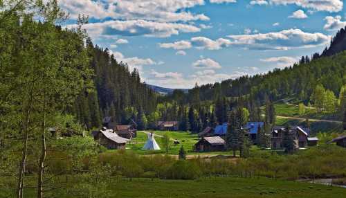 A scenic valley with a small village, teepee, and lush green trees under a blue sky with fluffy clouds.