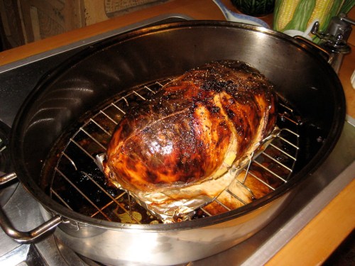 A large, roasted ham resting on a rack in a metal roasting pan, with a golden-brown, crispy exterior.