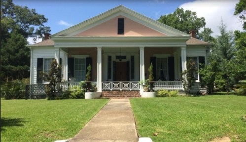 A charming two-story house with a pink facade, white columns, and a green lawn, surrounded by trees.