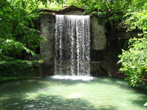 A serene waterfall cascading into a clear pool, surrounded by lush green trees and foliage.