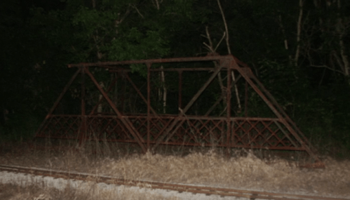 A rusted metal bridge structure stands in a dark, overgrown area near a railway track.