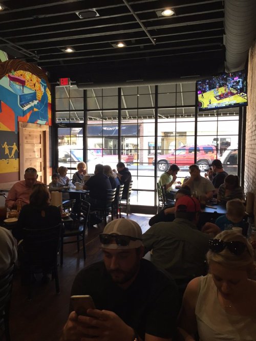 A busy restaurant interior with diners at tables, a TV showing a sports game, and large windows overlooking the street.