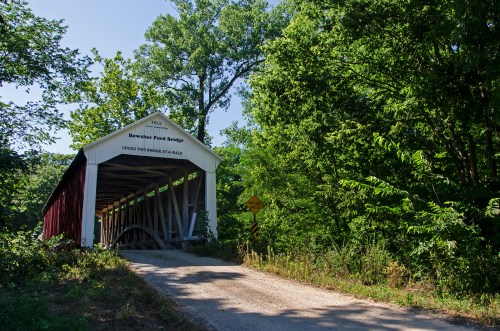 A red and white covered bridge surrounded by lush green trees, with a gravel road leading to it.