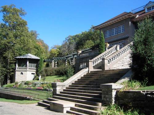 A grand stone staircase leads to a large, elegant house surrounded by lush greenery and a gazebo in the background.