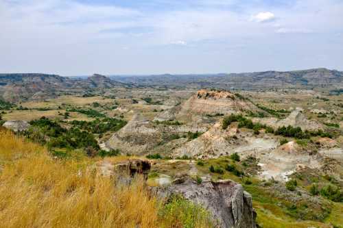 A panoramic view of rolling hills and valleys under a cloudy sky, with patches of grass and sparse vegetation.