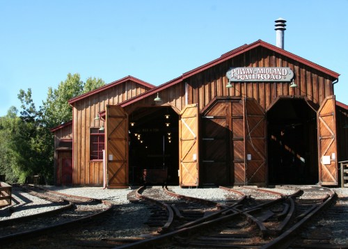 Historic wooden train depot with large doors, surrounded by railroad tracks and greenery. Clear blue sky above.