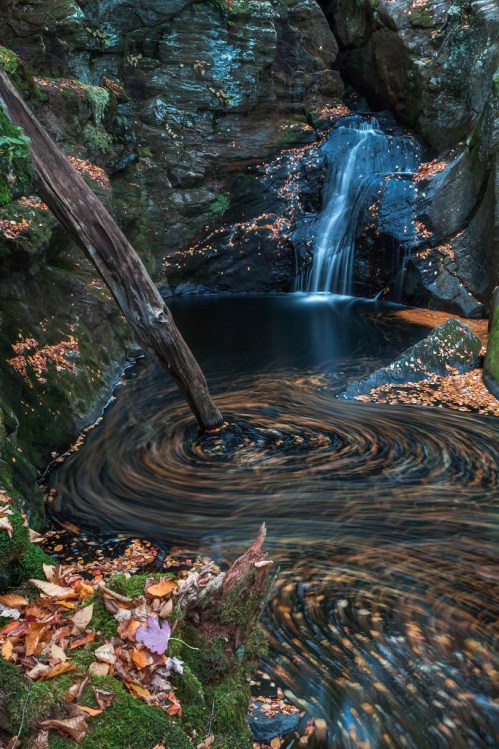 A serene waterfall cascades into a dark pool, surrounded by rocky cliffs and autumn leaves swirling in the water.