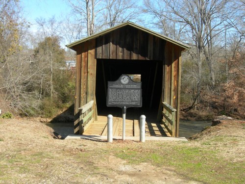 A wooden covered bridge with a historical marker, surrounded by trees and a grassy area.