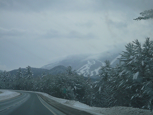 A winding road through a snowy landscape, with mountains and trees covered in white under a cloudy sky.