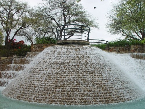 A cascading waterfall flows over a stone structure, surrounded by greenery and trees under a cloudy sky.