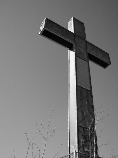 A tall, metallic cross stands against a clear sky, surrounded by bare branches in black and white.