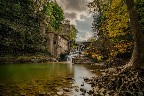 A serene waterfall cascades over rocks, surrounded by autumn foliage and an old stone building in a tranquil setting.