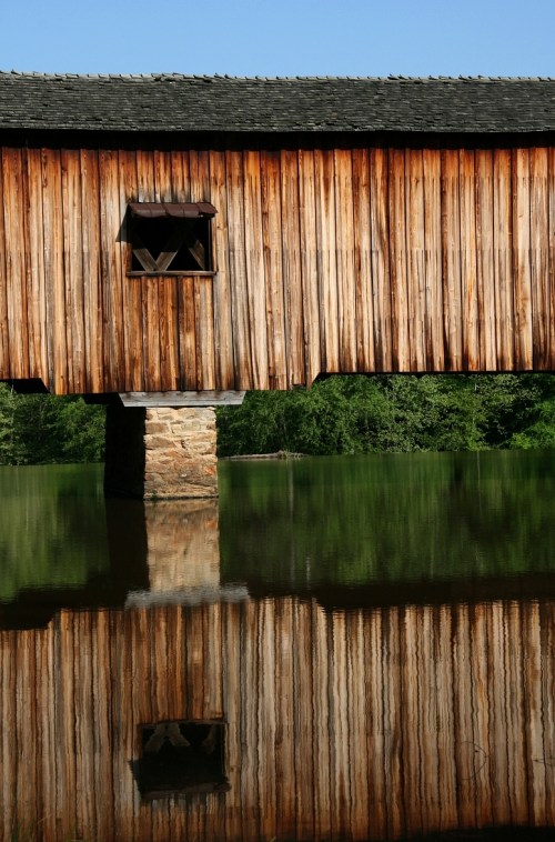 A wooden bridge with a stone support, reflecting in calm water surrounded by greenery.