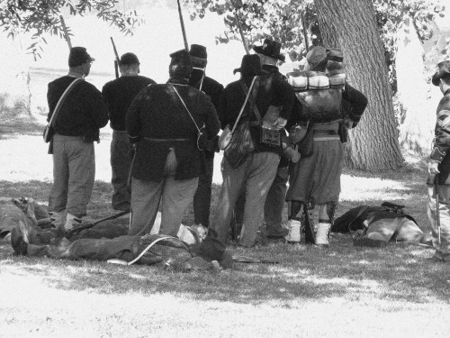 A group of soldiers in historical uniforms stands with rifles, some resting on the ground nearby, in a black-and-white scene.