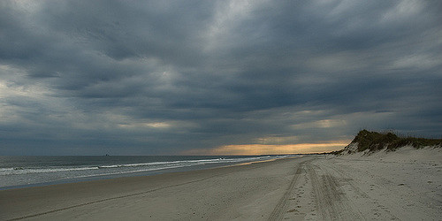 A serene beach scene with dark clouds overhead and a hint of sunset on the horizon, featuring soft sand and gentle waves.