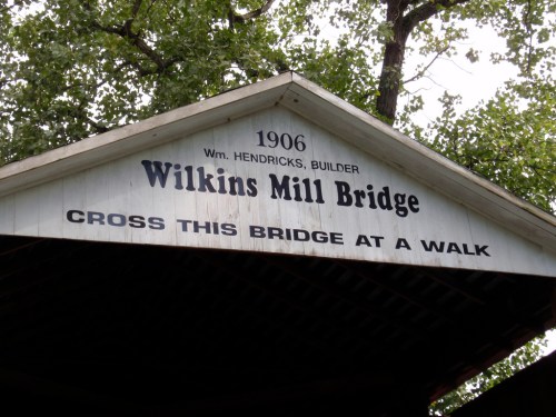 Sign on a covered bridge reads "Wilkins Mill Bridge, 1906, Wm. Hendricks, Builder. Cross this bridge at a walk."