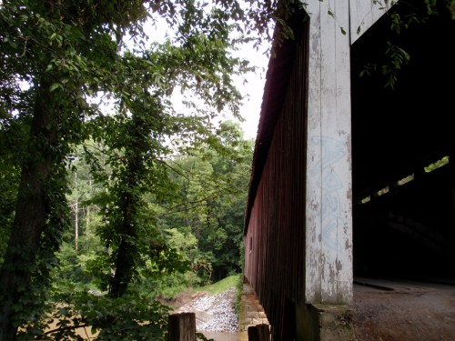 A covered bridge partially obscured by trees, with a riverbank visible in the background.