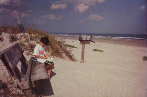 A boy sits on a bench by the beach, reading a book near a sign that says "Kindred Spirit." Ocean waves in the background.