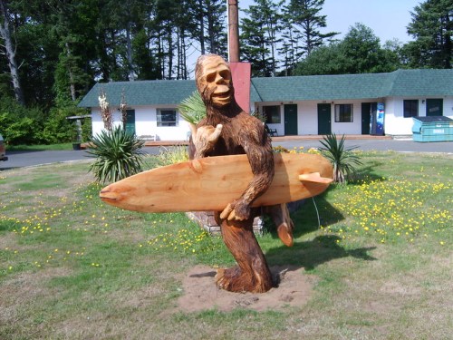 A wooden sculpture of a Bigfoot holding a surfboard, set in a grassy area near a building and trees.