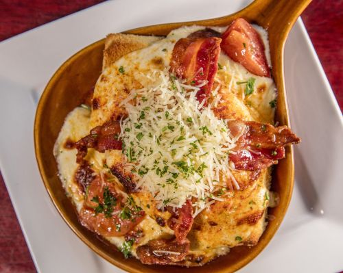 A baked dish with toast, cheese, bacon, tomatoes, and herbs, served in a brown bowl on a white plate.