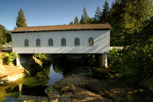 A white covered bridge spans a calm river, surrounded by lush green trees and a clear blue sky.