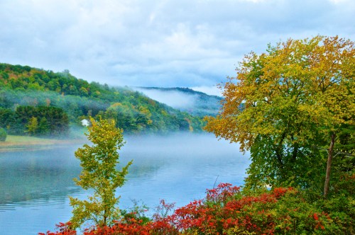 A serene lake surrounded by misty hills and colorful autumn trees under a cloudy sky.