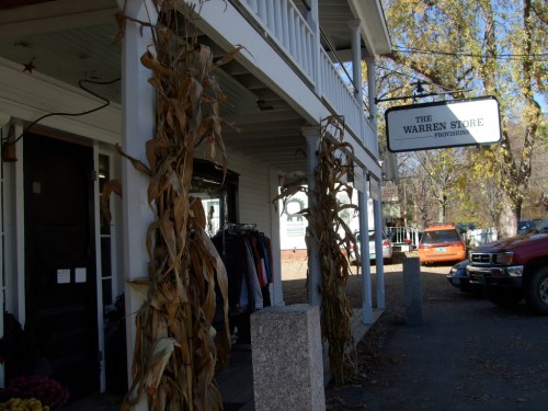 A quaint store front with a sign reading "The Warren Store," surrounded by autumn decorations and parked cars.