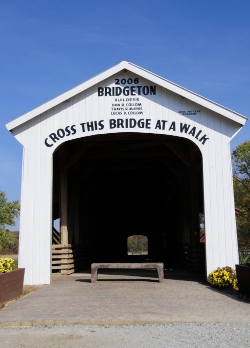 Covered bridge entrance with a sign reading "Cross This Bridge at a Walk" and the year 2006. Flowers in foreground.