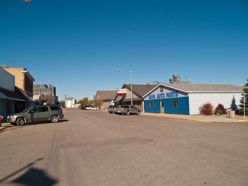 A quiet street scene featuring a blue auto parts store, parked cars, and clear blue skies.