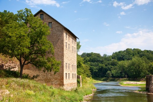 Historic stone building beside a river, surrounded by greenery and under a blue sky with scattered clouds.