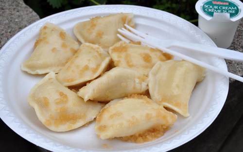 A plate of dumplings topped with caramelized onions, served with a side of sour cream. Two plastic forks are included.