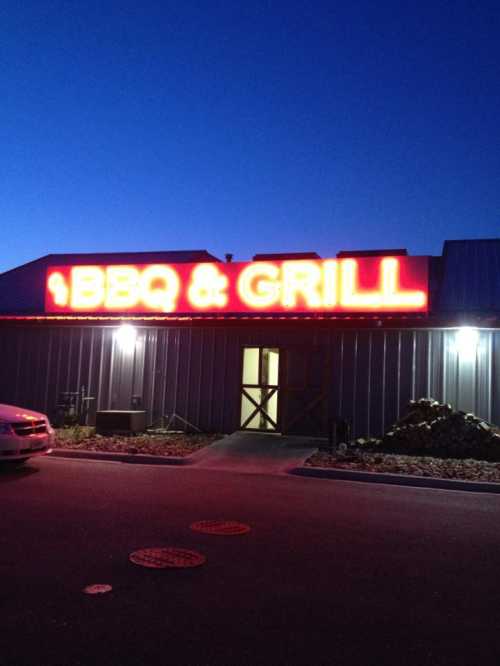 A brightly lit BBQ & Grill sign on a building at dusk, with a parking area in front.