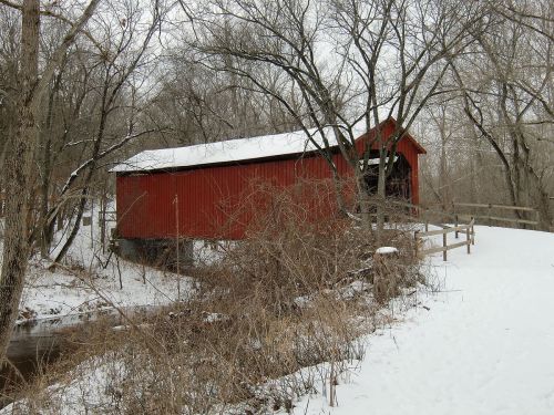 A red covered bridge surrounded by snow-covered trees and a creek, creating a serene winter landscape.