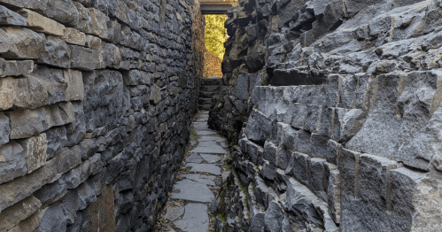 Narrow stone pathway between tall, rugged stone walls, leading to a bright opening at the end.