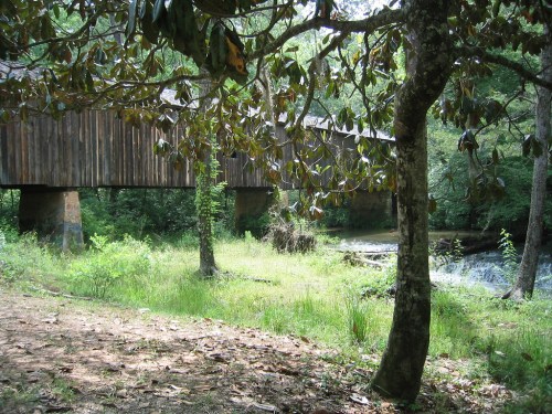 A wooden covered bridge spans a stream, surrounded by lush greenery and trees in a serene natural setting.