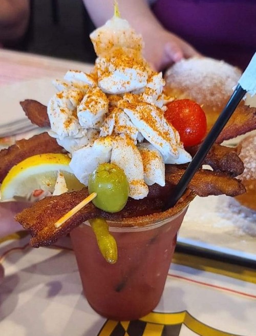 A colorful drink topped with whipped cream, bacon strips, lemon, cherry, and olives, served in a clear cup.